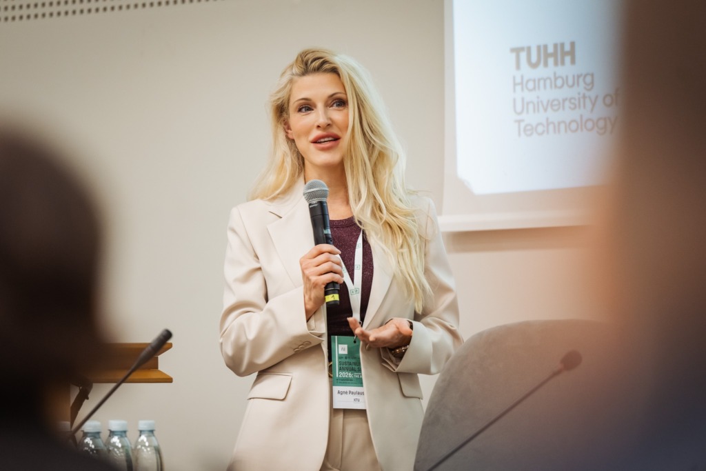 A speaker holds a microphone while presenting at the SustAInLivWork Annual Summit 2026 with Hamburg University of Technology displayed behind her. The event brings together AI expertise and perspectives across sectors.